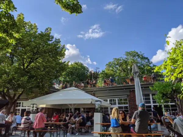 Menschen genießen das Wetter im Golgatha Biergarten. Sonnenschein, grüne Bäume und ein blauer Himmel bilden die Kulisse.