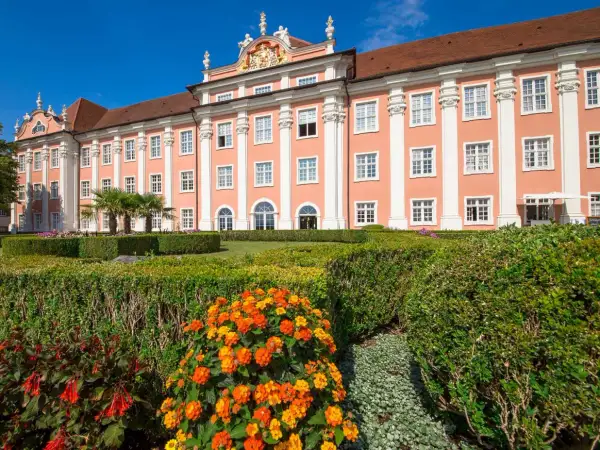 Das Meersburg Neues Schloss erstrahlt in Rosa vor blauem Himmel. Ein gepflegter Garten mit bunten Blumen und Palmen lädt zum Verweilen ein.